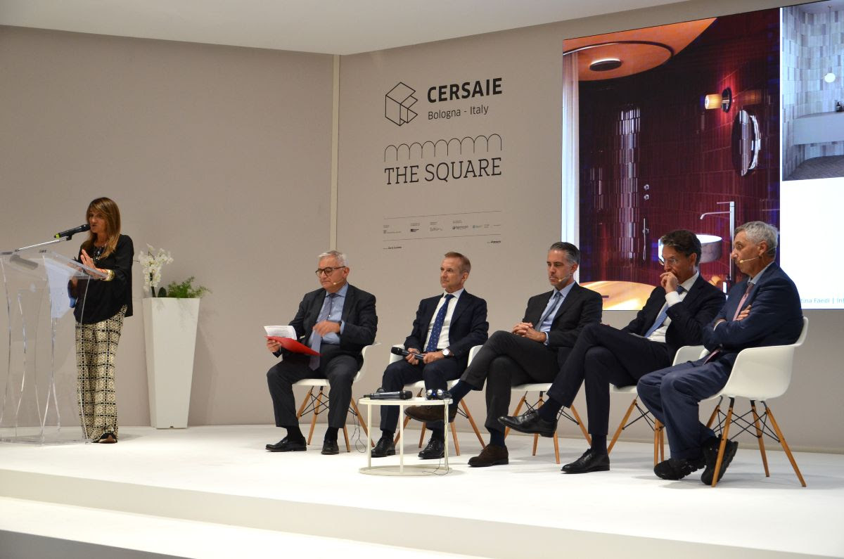 Image of 1 speaker presenting at a podium while the other speakers wait in a line of chairs on the stage