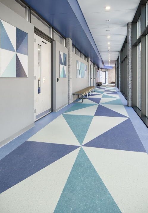 Hallway with doors on left and tall windows on right, with triangles of teal and bluish purple in VCT flooring, walls and ceiling